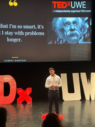 Image of PhD student Asaad Biqai on a stage in front of a presentation with an image of Albert Einstein and the quote: 'it's not that I'm so smart, it's just that I stay with problems longer'. 